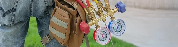 A technician, wearing jeans and a work bag, holds HVAC manifold gauges with red and blue pressure dials